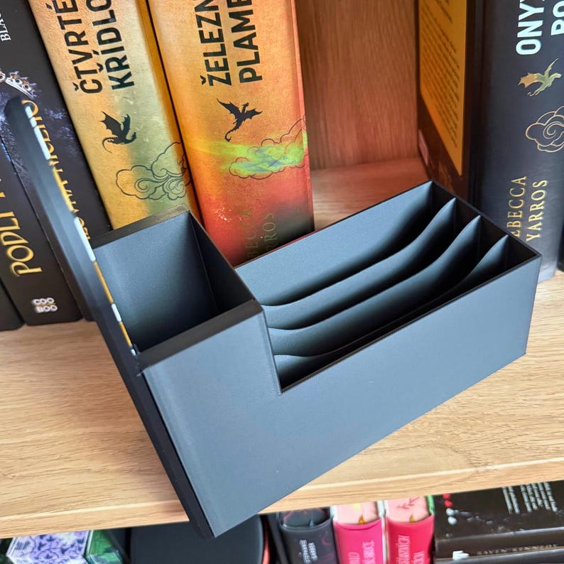 May include: A gray, multi-compartment organizer, likely for books or documents, sits on a wooden shelf. The organizer has a stepped design with several vertical dividers. Books with colorful spines are visible in the background.
