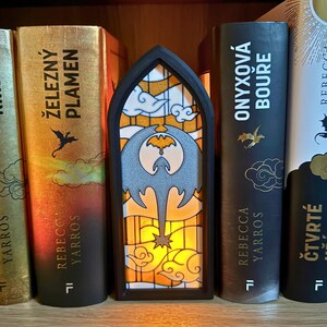 May include: A black framed stained glass night light with a dragon design. The stained glass features a gray dragon, orange and yellow clouds, and a star. The night light is placed between several books.