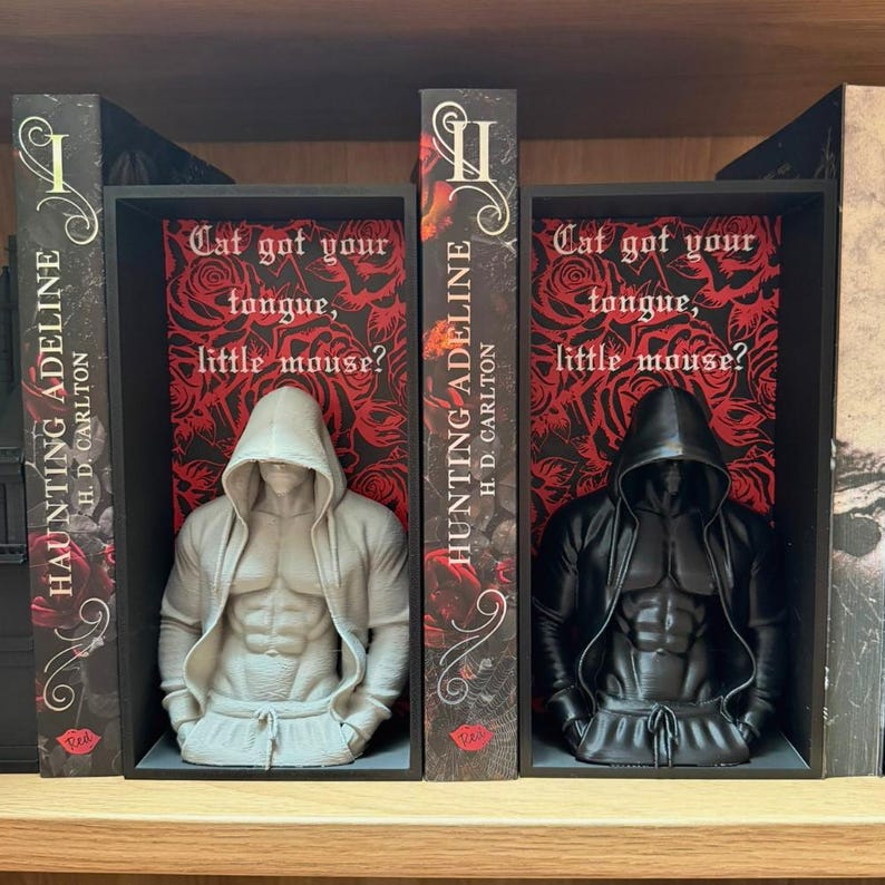 Puede incluir: Dos sujetalibros con figuras musculosas con capucha, flanqueando libros titulados "Haunting Adeline" de H.D. Carlton. Los sujetalibros est&aacute;n en una estanter&iacute;a negra con el texto "Cat got your tongue, little mouse?" sobre un fondo floral rojo.