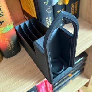 May include: A black 3D-printed book organizer with multiple compartments and a handle. The organizer is designed to hold books upright on a wooden shelf. The design includes a circular holder, possibly for a pen or small item.
