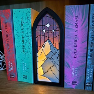 May include: A black framed, stained glass-style night light featuring a mountain scene with two stars. The light is placed between several books with colorful spines. The books are titled in a foreign language and are by Sarah J. Maas.