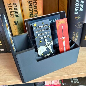 May include: A dark gray book organizer with a tablet and two bookmarks. One bookmark is red with a sword design, and the other is black with a dragon graphic and the text "A DRAGON WITHOUT ITS RIDER IS A TRAGEDY". Books are visible in the background.