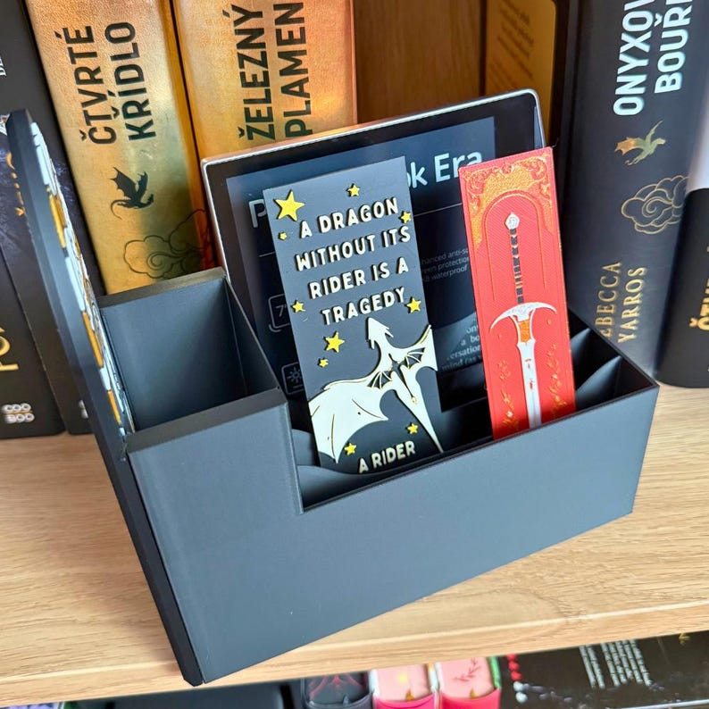 May include: A dark gray book organizer with a tablet and two bookmarks. One bookmark is red with a sword design, and the other is black with a dragon graphic and the text "A DRAGON WITHOUT ITS RIDER IS A TRAGEDY". Books are visible in the background.