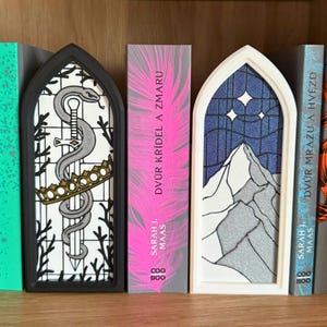 May include: Three decorative bookends in a bookshelf setting. One features a snake and sword design, another a mountain scene with stars, and the third a pink book spine with text. The bookends are in black and white frames.