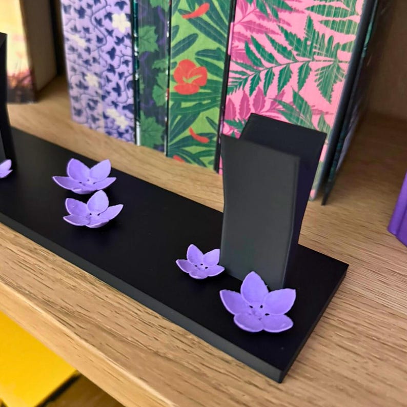 May include: A black wooden display stand with three upright supports and several small, purple flower-shaped decorations. The stand is on a wooden shelf, with colorful book spines in the background.