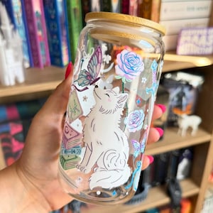 Op de afbeelding: Een doorzichtig glas met een bamboe deksel, voorzien van een witte vosillustratie, pastelkleurige vlinders, gestapelde boeken en blauwe rozen. Het ontwerp bevat fijne lijntekeningen en zachte roze, blauwe en groene accenten op het glas.