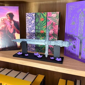 May include: A decorative silver sword with an ornate handle rests on a black stand, accented by purple flower-shaped decorations. The sword is displayed in front of a row of books with floral and illustrated covers, creating a fantasy theme.