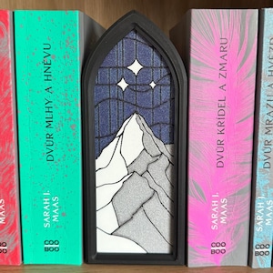 May include: A black, arched bookend featuring a mountain scene with a starry night sky. The bookend is placed between books with titles in a foreign language. The books have pink, green, and red covers.