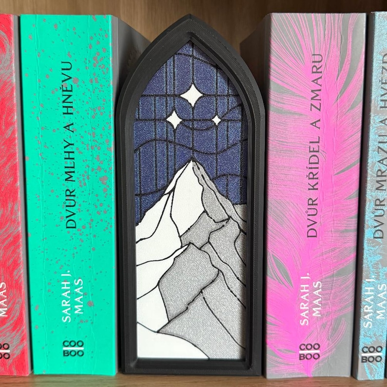 May include: A black, arched bookend featuring a mountain scene with a starry night sky. The bookend is placed between books with titles in a foreign language. The books have pink, green, and red covers.