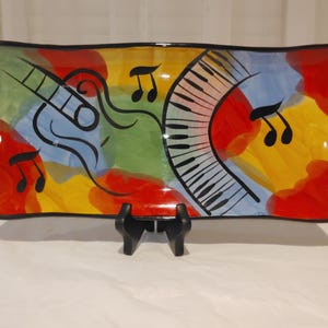 May include: A rectangular ceramic serving platter with a musical theme. The platter features a colorful abstract design with a guitar, piano keys, and musical notes. The colors include red, yellow, blue, and green, with black accents. The platter is displayed on a black stand.