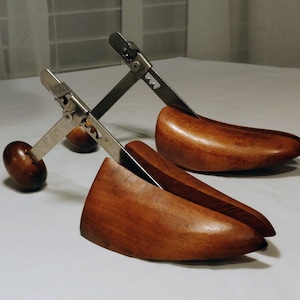 May include: A pair of wooden shoe trees featuring smooth, polished brown wood toe sections and adjustable metal heel mechanisms with rounded wooden knobs. The metal components include a notched tension bar for length adjustment.