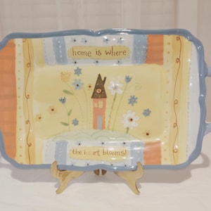 May include: Rectangular ceramic serving tray with a light blue border and handles. The tray features a painted design with a house, flowers, and the text "home is where" and "the heart blooms!". The tray is supported by a wooden stand.