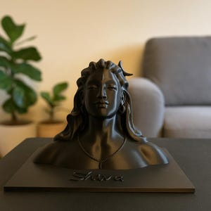 Könnte beinhalten: Eine schwarze Skulptur einer Gottheit mit langem Haar, Halskette und geschlossenen Augen. Die Skulptur ist auf einem schwarzen Sockel mit dem Namen "Shiva" angebracht. Der Hintergrund ist verschwommen und zeigt eine Wohnzimmereinrichtung.