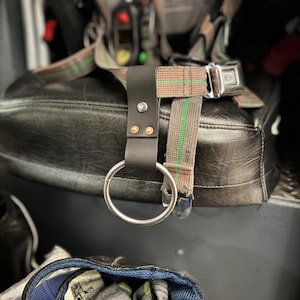 May include: A black leather strap with a silver metal ring attached to a seat. The strap is secured with rivets. Brown and green striped webbing is attached to a metal buckle. A hat with the text "10M" is in the foreground.