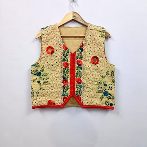 Puede incluir: Un chaleco sin mangas de tela beige claro con bordados intrincados de motivos florales circulares rojos, enredaderas verdes y detalles en azul. La prenda tiene un cuello en V, un borde rojo brillante en la parte delantera y un patrón festoneado.