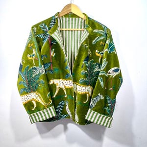 Puede incluir: Chaqueta de terciopelo verde con estampado de jungla con leopardos, cebras y pájaros. La chaqueta tiene forro y puños a rayas. Cuelga de una percha de madera.