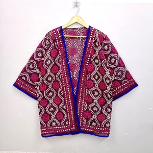 Könnte beinhalten: Eine transparente, magentafarbene Kimono-Jacke mit aufwendigen weißen und dunkelrosa Kreisstickereien, geometrischen Mustern und kleinen reflektierenden Spiegelakzenten. Das Kleidungsstück ist mit einem leuchtend blauen Saum an der Vorderseite und den Ärmeln versehen.