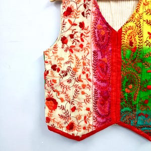 May include: A sleeveless vest featuring a vibrant color-blocked design with sections of cream, pink, yellow, and green. The garment is adorned with intricate red and orange floral and paisley embroidery throughout, finished with a solid red trim.