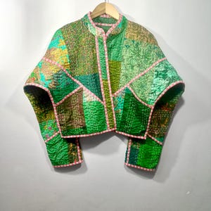 Peut inclure: Veste patchwork aux tons verts, marron et jaunes, avec des bordures roses. La veste est composée de divers morceaux de tissu cousus ensemble, créant un design unique. Elle a un col rond et des manches courtes, suspendue à un cintre en bois.