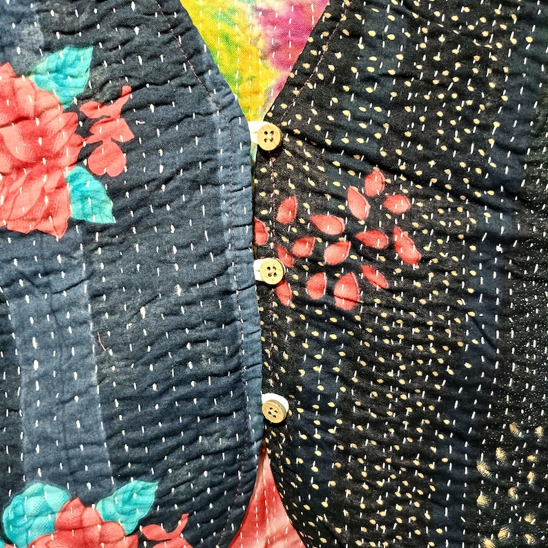 Op de afbeelding: Close-up van een gewatteerd textiel met een bloemmotief. De stof is donkerblauw met rode bloemen en blauwgroene bladeren. De rechterkant heeft een stippenpatroon met rode bladvormen en drie witte knopen.