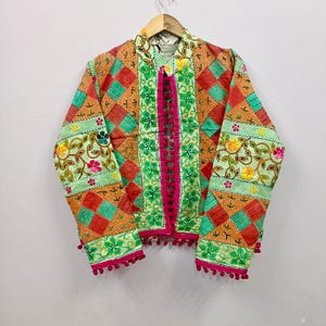 May include: A quilted jacket featuring a patchwork design of orange and teal diamond patterns, accented with light green floral embroidery panels. The front opening and cuffs are trimmed with bright pink fabric and small pink pom-pom fringe.