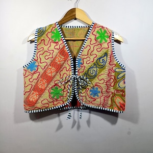 Può includere: Gilet ricamato colorato con chiusura a fiocco. Il tessuto beige è decorato con motivi floreali e astratti rossi, arancioni, verdi e blu. I bordi sono rifiniti con tessuto a righe blu navy e bianco. Il gilet è appeso.