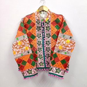 Op de afbeelding: Een jas met lange mouwen voorzien van een patchworkontwerp van oranje en groene vierkanten, geaccentueerd met bloemenborduursels in geel en groen. De sluiting aan de voorzijde bevat een rij kleine roze pompons, met extra roze pompons langs de zoom.