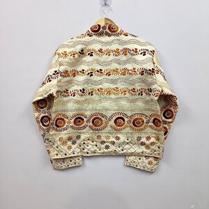 May include: A cream-colored jacket featuring intricate embroidery in shades of orange, brown, and gold. The design includes horizontal wavy lines, circular floral motifs, and a bottom border with small star patterns and reflective mirror accents.