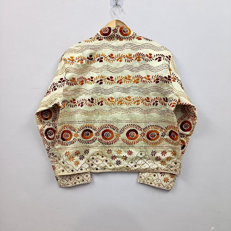 May include: A cream-colored jacket featuring intricate embroidery in shades of orange, brown, and gold. The design includes horizontal wavy lines, circular floral motifs, and a bottom border with small star patterns and reflective mirror accents.