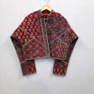 May include: A quilted jacket featuring a deep red base with intricate floral and geometric block-print patterns in blue and white. The garment has a high collar, long sleeves, and distinct black and white striped trim along the edges and seams.