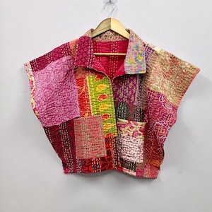Può includere: Giacca patchwork con una varietà di colori e motivi, tra cui rosa, rosso, verde e oro. La giacca ha un colletto, maniche corte e due tasche. È appesa a una gruccia di legno su sfondo bianco.