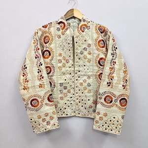 Könnte beinhalten: Eine cremefarbene Jacke mit aufwendigen Stickereien in Orange-, Braun- und Goldtönen. Das Kleidungsstück ist mit kleinen runden Spiegeln an den Vorderteilen und Ärmeln verziert, die in geometrischen und floralen Mustern angeordnet sind.