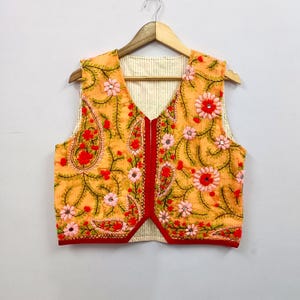 Può includere: Un gilet arancione ricamato con un motivo floreale. Il gilet ha uno scollo a V ed è decorato con fiori rossi e rosa. I bordi sono rifiniti con tessuto rosso. Il gilet è appeso a una gruccia di legno.