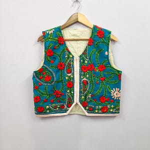 Könnte beinhalten: Eine türkisfarbene Weste mit roten Blumen und grünen Blättern bestickt. Die Weste hat einen V-Ausschnitt und ein cremefarbenes Futter. Das florale Design ist detailliert und bedeckt die Vorder- und Rückseite. Die Weste hängt an einem Holzbügel.