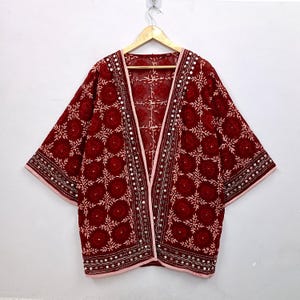 Könnte beinhalten: Eine dunkelrote, vorne offene Kimono-Jacke mit aufwendiger weißer Blumenstickerei und kleinen runden Spiegelverzierungen an den Vorderkanten und Ärmeln. Das Kleidungsstück hat weite Dreiviertelärmel und einen hellrosa Saum.
