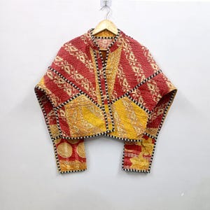Op de afbeelding: Een gewatteerd jasje met een patchworkontwerp in rood- en geeltinten met ingewikkelde patronen. Het kledingstuk heeft lange mouwen, een opstaande kraag en een zwart-wit geblokte bies langs de randen en naden, hangend aan een kledinghanger.