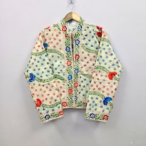 Puede incluir: Una chaqueta acolchada de color crema con un patrón integral de motivos florales y enredaderas bordadas en rojo, azul y verde. La prenda tiene mangas largas, un dobladillo recto y una abertura central en la parte delantera, todo con costuras.