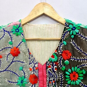 K&ouml;nnte beinhalten: Ein Oberteil mit V-Ausschnitt und geteiltem Design, bestehend aus einer hellgrauen und einer dunkelgrauen Seite, beide verziert mit leuchtender Blumenstickerei in Hellgr&uuml;n, Orange und Blau. Das Kleidungsst&uuml;ck hat eine mittige rosa Zierleiste.