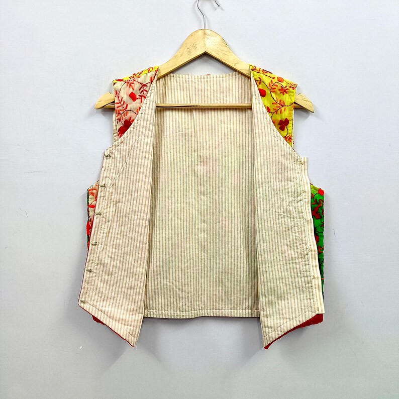 May include: A sleeveless vest featuring a beige and white vertical striped interior lining. The exterior panels are crafted from vibrant, multi-colored fabric with intricate floral embroidery in shades of red, yellow, and green, hanging on a hanger.