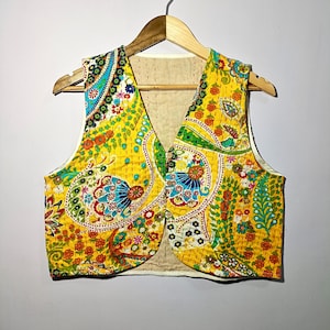 May include: A yellow vest with a colorful paisley pattern, featuring shades of blue, green, red, and orange. The vest is button-down and has a V-neck. It is hanging on a wooden hanger.