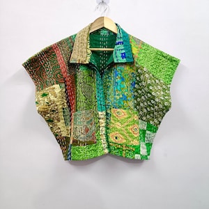 Puede incluir: Una chaqueta patchwork en tonos verdes, rojos y beige cuelga de una percha de madera. La chaqueta presenta un cuello, mangas cortas y una parte delantera abotonada. La tela es una mezcla de piezas estampadas y lisas.