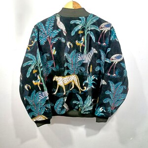 May include: Black bomber jacket with a jungle-themed pattern. The design features a leopard, zebra, flamingo, and parrot amidst lush foliage in teal, yellow, and white. The jacket has a dark green collar and cuffs.