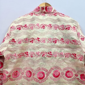 May include: An off-white jacket featuring intricate pink floral embroidery. The design includes horizontal wavy lines, repeating leaf patterns, and circular flower motifs in various shades of pink, all stitched onto a light-colored fabric base.