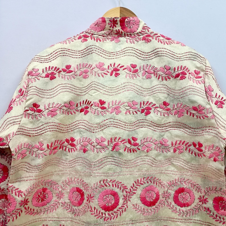 May include: An off-white jacket featuring intricate pink floral embroidery. The design includes horizontal wavy lines, repeating leaf patterns, and circular flower motifs in various shades of pink, all stitched onto a light-colored fabric base.