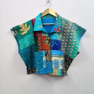 May include: A patchwork top in shades of teal, blue, and red, with a collared neckline and short sleeves. The top is made from various fabric pieces, stitched together with visible seams. The garment is hanging on a wooden hanger against a white background.