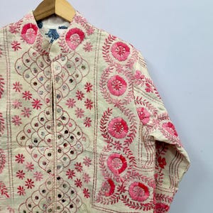 May include: An off-white long-sleeve jacket featuring intricate pink floral embroidery and small circular mirrors arranged in a diamond pattern. The garment has a mandarin collar and is displayed on a wooden hanger against a plain background.