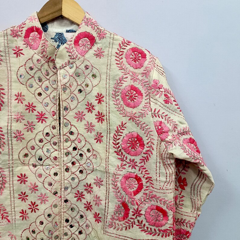May include: An off-white long-sleeve jacket featuring intricate pink floral embroidery and small circular mirrors arranged in a diamond pattern. The garment has a mandarin collar and is displayed on a wooden hanger against a plain background.