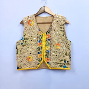 Puede incluir: Chaleco beige bordado con diseño de cuello en V. El chaleco presenta bordados florales coloridos en naranja, amarillo y azul, con pequeños detalles de espejo. Los bordes están ribeteados con tela amarilla. El chaleco se muestra en una percha de madera.