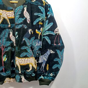 May include: A black bomber jacket with a vibrant jungle-themed print. The design features leopards, zebras, toucans, and flamingos amidst lush foliage in shades of green, blue, and yellow. The jacket has a zip-up front and ribbed cuffs.