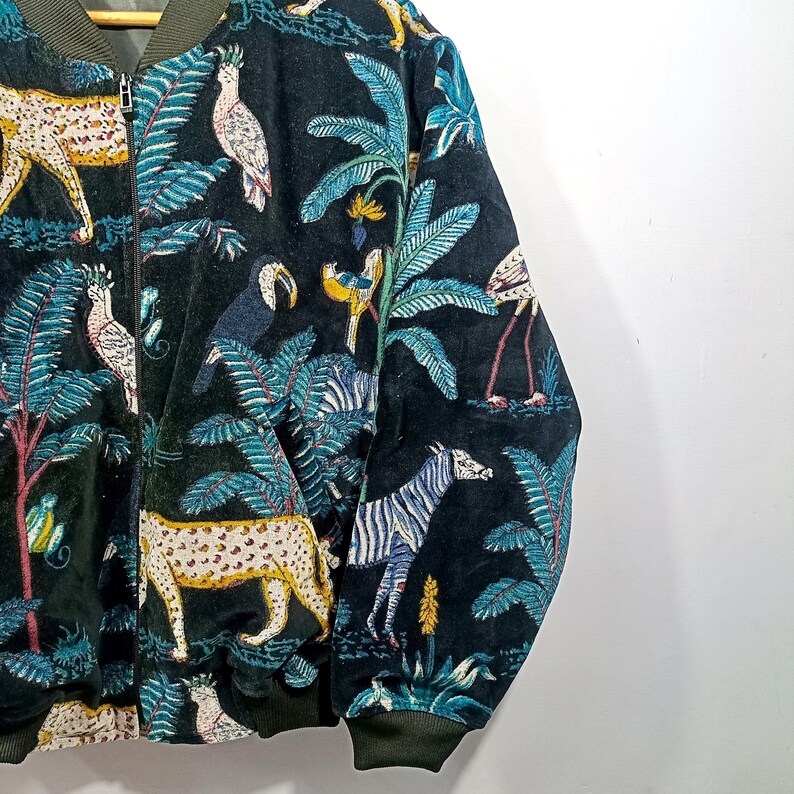 May include: A black bomber jacket with a vibrant jungle-themed print. The design features leopards, zebras, toucans, and flamingos amidst lush foliage in shades of green, blue, and yellow. The jacket has a zip-up front and ribbed cuffs.